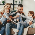 Couple sits on a couch, laughing while holding their two young children on their laps.